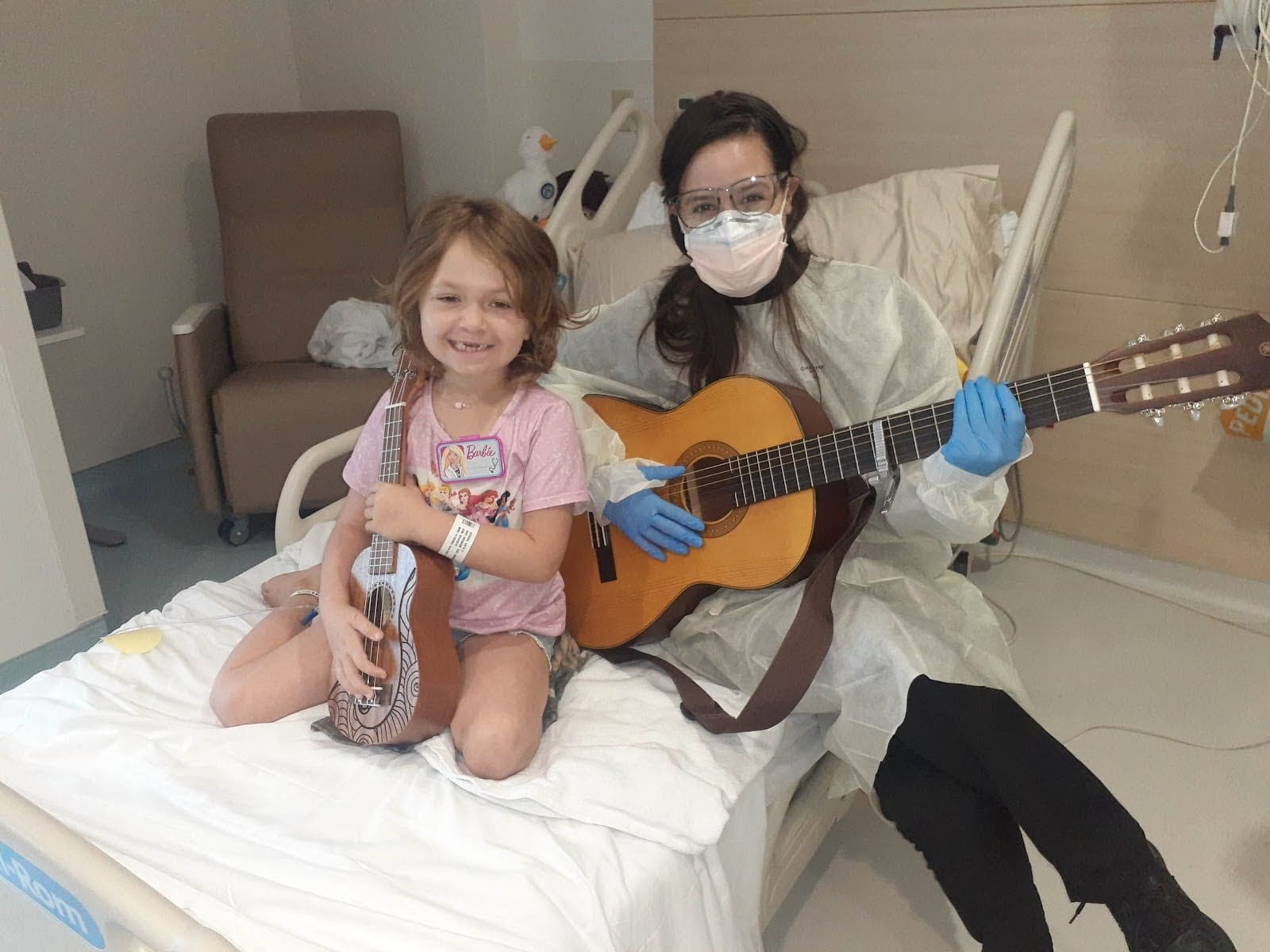 Child in a hospital bed smiling and holding her ukulele in a hospital bed along with her music therapist who is holding a guitar