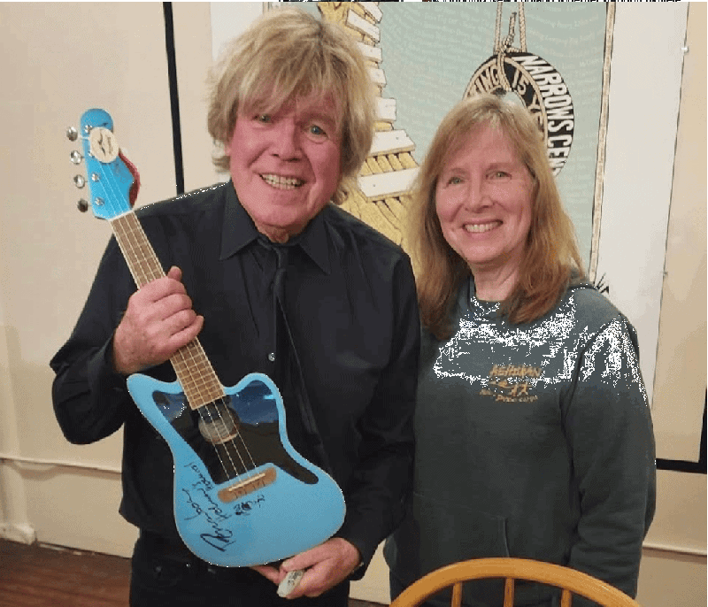 A smiling Peter Noone of Herman’s Hermits holding a signed light blue acoustic-electric ukulele, standing next to a smiling woman